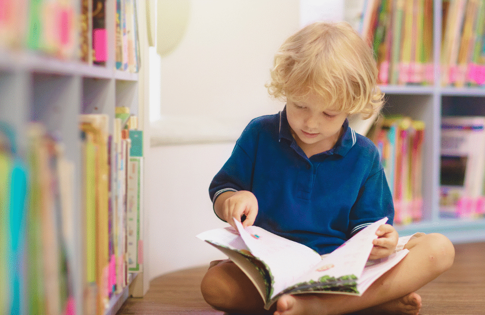 Criança sentada lendo sozinha perto de estante de livros, simbolizando dificuldades de comunicação e aprendizagem relacionadas ao TPAC