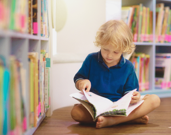 Criança sentada lendo sozinha perto de estante de livros, simbolizando dificuldades de comunicação e aprendizagem relacionadas ao TPAC
